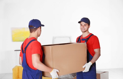 Two male removal workers dressed in red shirts, blue overalls, and blue caps are carrying a large cardboard box together in a bright, minimalist indoor setting. One worker faces the camera with a smile, while the other has his back to the camera. Both are wearing white gloves to handle the box safely. The background features a white wall with minimal decoration, including a small abstract painting with yellow and white tones, and a glimpse of a yellow piece of furniture on the left. The scene suggests a professional house removal or relocation service, with a focus on careful handling of household belongings. The lighting is natural and even, highlighting the textures of the cardboard box and the workers' clothing. The image emphasizes teamwork and professionalism in a residential or office moving context, subtly referencing services available in the Sutton area as part of Sutton Man with Van's offerings in the removals sector.