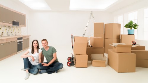 A young couple sitting on the floor of a bright, modern kitchen and living area with large windows, surrounded by unpacked cardboard boxes and packing materials indicative of a recent or ongoing house move. The woman has long dark hair and is dressed in a white top and jeans, while the man wears a green t-shirt and jeans, both smiling toward the camera. In the background, there's a kitchen with light-colored cabinets, a tiled backsplash, and built-in appliances. To the right, stacked boxes of various sizes are placed on the floor, some open with flaps folded back, and there is a red and black hand truck near the boxes. A tall, step ladder leans against the wall near the window, and a small potted plant with green leaves sits on a windowsill, adding a touch of greenery to the space. The room is filled with natural daylight, creating a welcoming environment that reflects a domestic setting suitable for a home removal service in Sutton or surrounding areas of south London. This scene illustrates a house clearance or relocation process, embodying the activities of Sutton Man with Van's removals services.